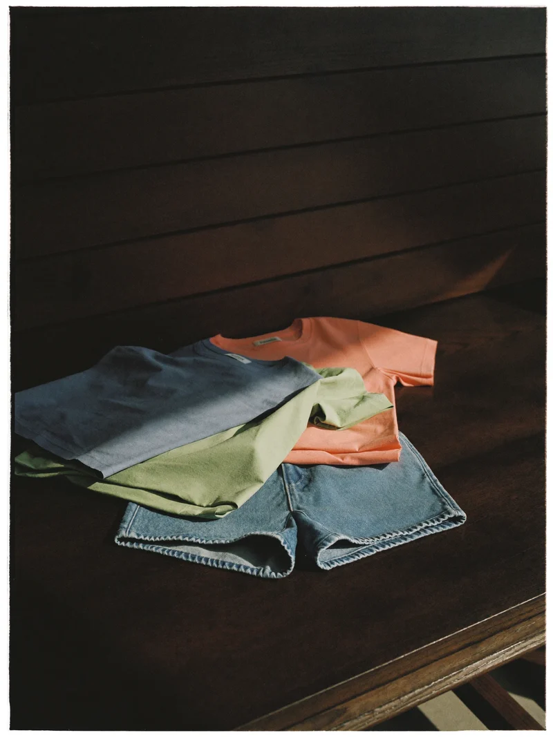 Tricouri simple și pantaloni scurți din denim în pasteluri stinse, pe o masă de lemn.